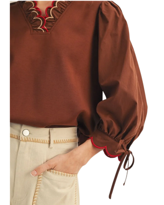 Women’s chocolate brown blouse with red scalloped trim, decorative V-neckline, puff sleeves and tie cuffs, styled with cream pants.