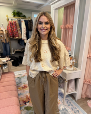 Woman wearing an oatmeal oversized short sleeve knit sweater with blush pink horseshoe accents, ribbed neckline, and cuffs, styled with taupe pants.