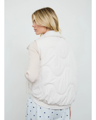 Women's ivory quilted puffer vest with high collar, front pockets and adjustable side tie details styled over a cream long-sleeve top.
