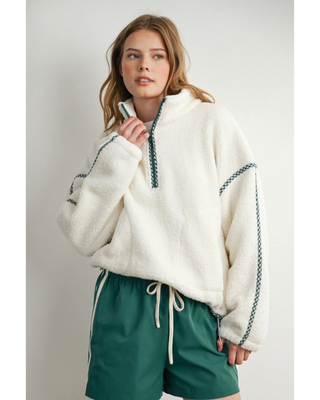 A woman wearing a cream sherpa pullover with dark green braided trim and matching green drawstring shorts, photographed against a plain light gray studio background.