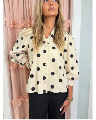Women's ivory button-front blouse with black polka dots, a ruffle collar, long sleeves, and a relaxed fit, styled for a chic boutique outfit.
