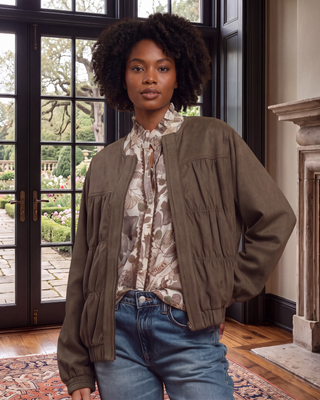 A woman wearing a brownish-olive utility jacket over a patterned blouse and blue jeans, standing in a room with large black-framed glass doors and a fireplace.