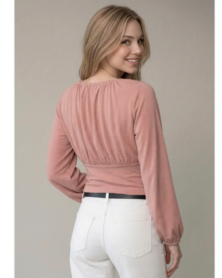 A woman shown from the back wearing a dusty rose long-sleeve top and white pants, standing against a plain beige studio background.