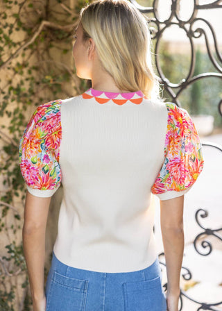 Woman wearing a white top with colorful floral sleeves and blue pants, standing against a decorative metal railing.