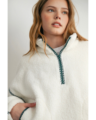 A woman wearing a white sherpa fleece pullover with a half-zip neckline and dark green braided trim, shown against a light gray studio background.