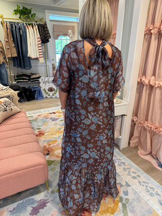 Woman wearing a chocolate brown maxi dress with dusty blue floral print, smocked mock neckline, short flutter sleeves, relaxed fit, and ruffled hem, styled with tan boots.