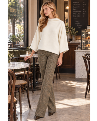 A woman stands in a cafe wearing a cream-colored knit mock-neck top and brown and beige houndstooth patterned pants.