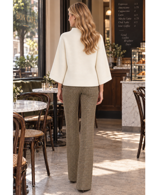 A woman stands with her back to the camera inside a cafe, wearing a cream-colored wide-sleeve mock-neck top and brown and beige houndstooth trousers.