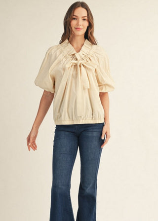 A woman modeling a cream-colored short-sleeve blouse with a ruffled tie neckline, paired with dark blue flared jeans and brown shoes, against a solid light beige background.