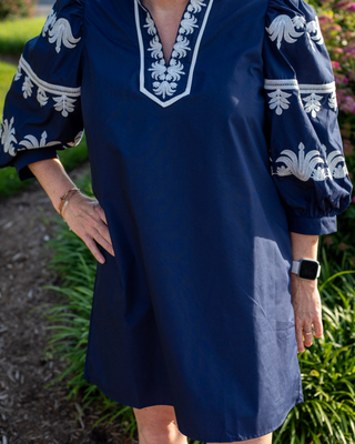 A navy blue tunic dress with white embroidered floral patterns on the sleeves and V-neckline, worn by a model in an outdoor garden setting.