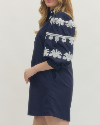 A woman wearing a navy blue short dress with white embroidered patterns on the shoulders and upper chest, shown from a side profile against a light gray studio background.