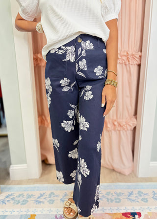 Woman wearing navy blue wide leg pants with a white floral print, front patch pockets, and a high-rise waist paired with a white textured top.