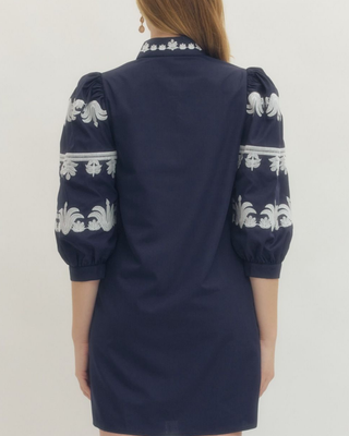 A woman viewed from the back wearing a navy blue dress with white floral embroidery on the sleeves and collar.