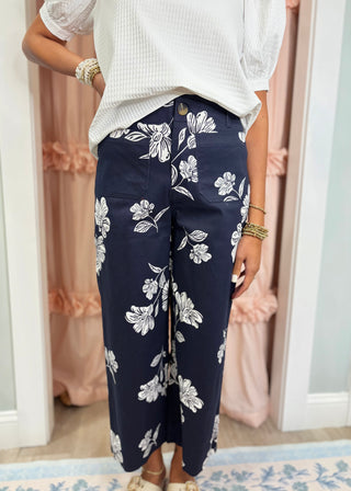 Woman wearing navy blue wide leg pants with a white floral print, front patch pockets, and a high-rise waist paired with a white textured top.
