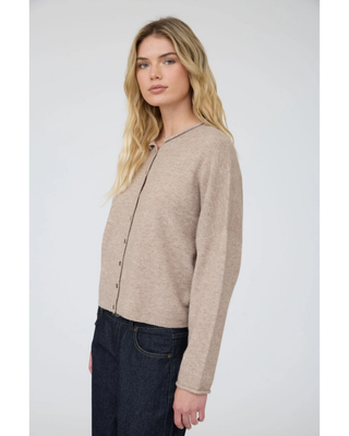 A blonde woman wearing a light beige knit cardigan with a buttoned front and dark blue jeans, photographed against a neutral light gray studio background.