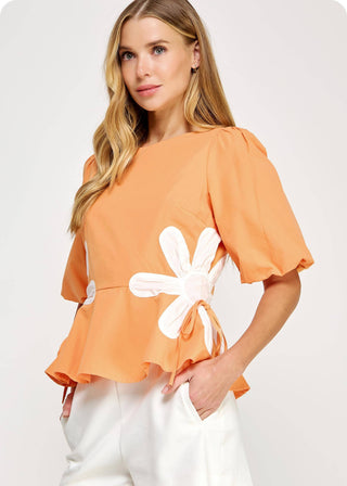 Woman wearing an orange top with a white flower design and white shorts on a light gray background