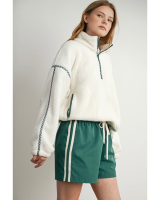 A woman modeling a cream sherpa half-zip pullover with green braided trim and green shorts with white side stripes, photographed against a light gray studio background.