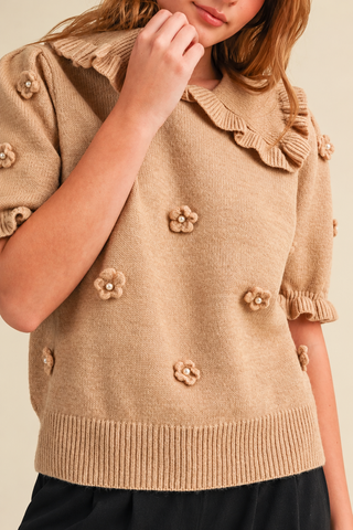 Model wearing a camel beige women's knit sweater with a ruffled collar, short puff sleeves, pearl-centered 3D floral appliqués, and ribbed hem, styled with black pants.