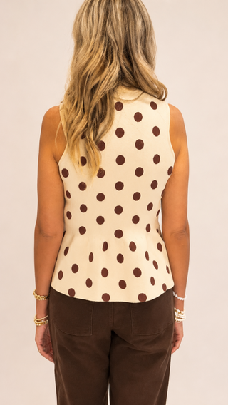 Woman wearing a cream sleeveless mock neck peplum sweater tank with chocolate brown polka dots paired with brown tailored pants against a neutral studio background.