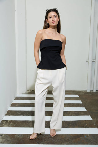 Model wearing a black strapless peplum top with a fitted bodice and flared hem paired with white tailored pants.