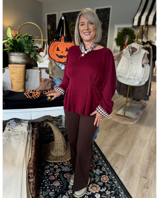 Woman wearing a burgundy long sleeve peplum top with contrasting brown and white gingham cuffs, a round neckline, and flowy hem, styled with brown pants.