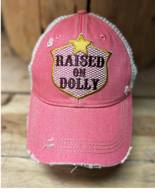 Pink distressed mesh trucker hat with embroidered "Raised on Dolly" patch, yellow star detail, and adjustable snapback closure.