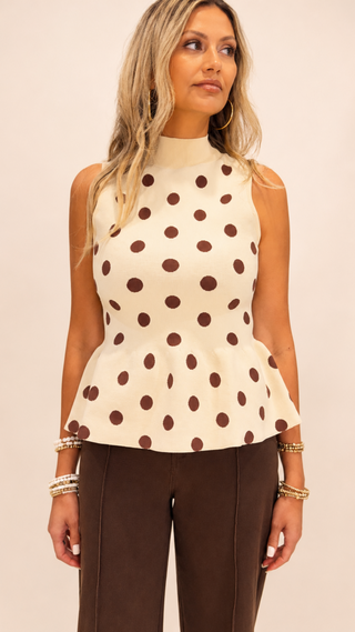 Woman wearing a cream sleeveless mock neck peplum sweater tank with chocolate brown polka dots paired with brown tailored pants against a neutral studio background.