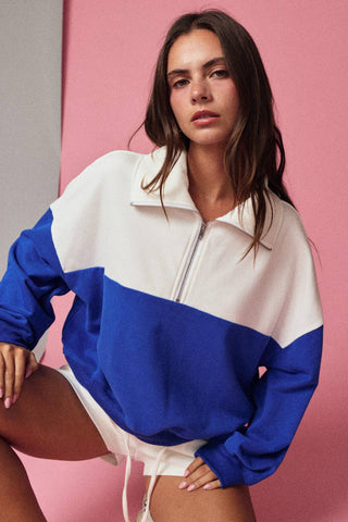 Woman wearing an oversized blue and white color-block quarter-zip pullover sweatshirt with white shorts against a pink background.