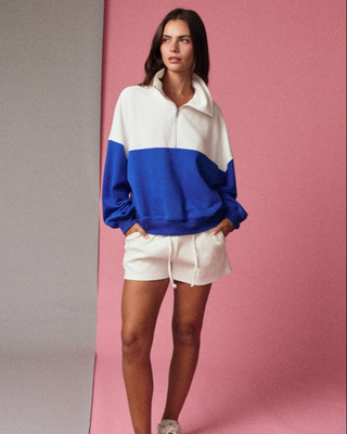 Woman wearing an oversized blue and white color-block quarter-zip pullover sweatshirt with white shorts against a pink background.