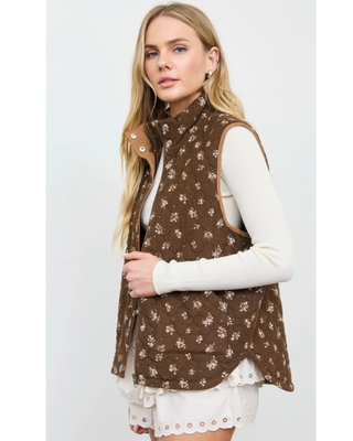 Woman wearing a chocolate brown quilted vest with a delicate floral print, stand-up collar, snap-button and zipper closure, and front patch pockets over a cream ribbed top.