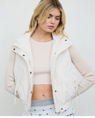 Women's ivory quilted puffer vest with high collar, front pockets and adjustable side tie details styled over a cream long-sleeve top.