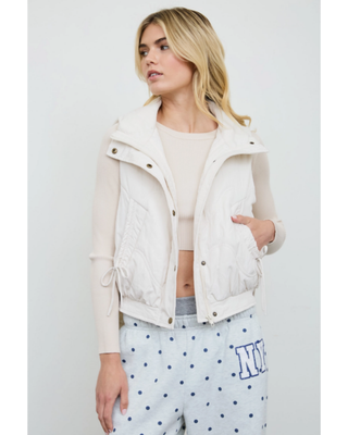 Women's ivory quilted puffer vest with high collar, front pockets and adjustable side tie details styled over a cream long-sleeve top.