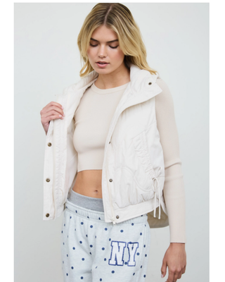 Women's ivory quilted puffer vest with high collar, front pockets and adjustable side tie details styled over a cream long-sleeve top.