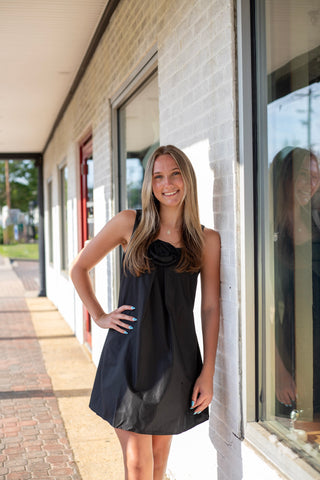 Model wearing a sleeveless black swing dress with a statement rosette neckline and relaxed above-the-knee silhouette.