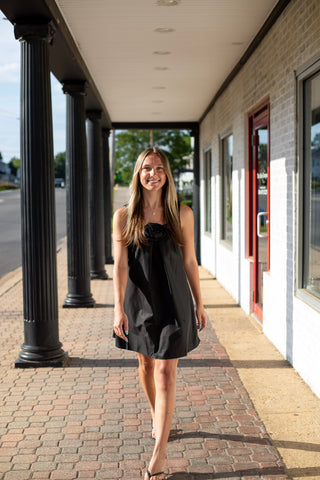 Model wearing a sleeveless black swing dress with a statement rosette neckline and relaxed above-the-knee silhouette.