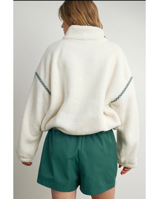 A person shown from the back wearing a cream sherpa fleece pullover with dark green braided stripe accents on the sleeves and teal-green shorts, against a plain light gray studio background.
