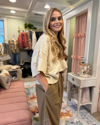 Woman wearing an oatmeal oversized short sleeve knit sweater with blush pink horseshoe accents, ribbed neckline, and cuffs, styled with taupe pants.