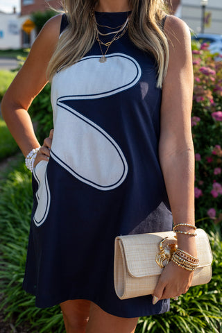 A woman wearing a sleeveless navy blue dress with a large white abstract graphic on the front, accessorized with layered gold necklaces, bracelets, and a beige clutch bag, set against a blurred outdoor garden background.