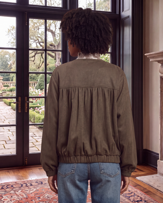 A person viewed from behind wearing an olive brown gathered bomber jacket and blue jeans, standing in a room with large black-framed glass doors looking out onto a garden.