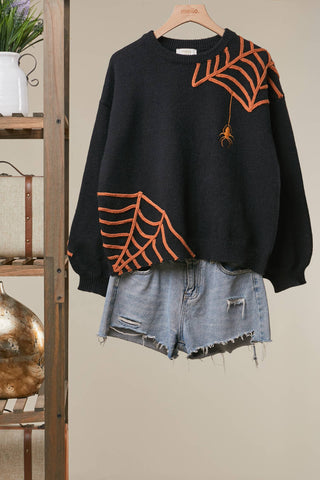 Black Halloween knit sweater with orange embroidered spiderweb designs and hanging spider detail, styled with distressed denim shorts.