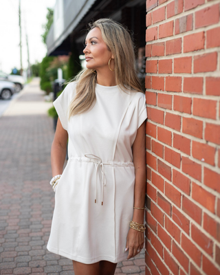 Model wearing an ivory drawstring shift mini dress with cap sleeves, side pockets, and a crew neckline.