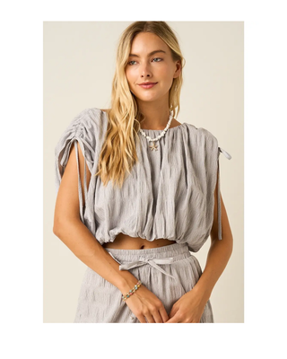 The Maren Bubble Top in light gray featuring textured fabric, adjustable shoulder ties, an elastic neckline, and a flattering bubble hem.