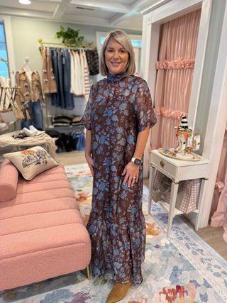 Woman wearing a chocolate brown maxi dress with dusty blue floral print, smocked mock neckline, short flutter sleeves, relaxed fit, and ruffled hem, styled with tan boots.