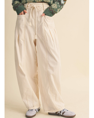 A person wearing high-waisted, wide-leg cream pants with a drawstring waist, paired with a green patterned sweater and gray sneakers, set against a solid beige background.