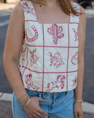 Model wearing an ivory sleeveless crop top with red embroidered western patchwork featuring cowboy boots, cacti, horseshoes, roses, and a square neckline.