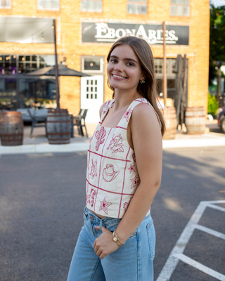 Model wearing an ivory sleeveless crop top with red embroidered western patchwork featuring cowboy boots, cacti, horseshoes, roses, and a square neckline.