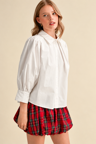 Model wearing a white button-down blouse with puff sleeves, pointed collar, and wide cuffs, paired with a red plaid bubble skirt.