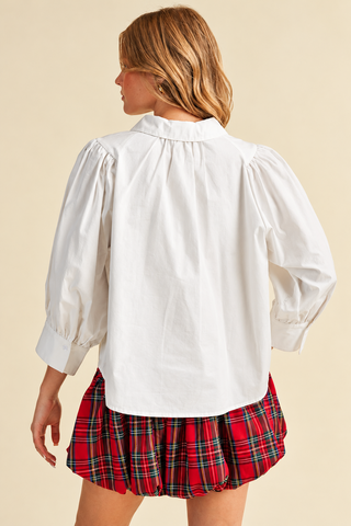 Model wearing a white button-down blouse with puff sleeves, pointed collar, and wide cuffs, paired with a red plaid bubble skirt.