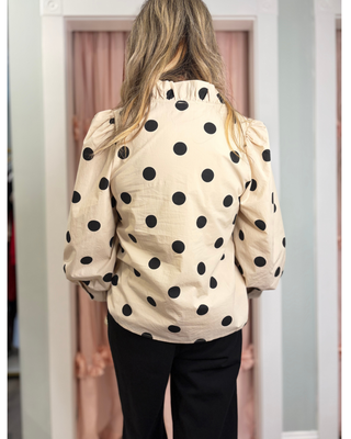 Women's ivory button-front blouse with black polka dots, a ruffle collar, long sleeves, and a relaxed fit, styled for a chic boutique outfit.