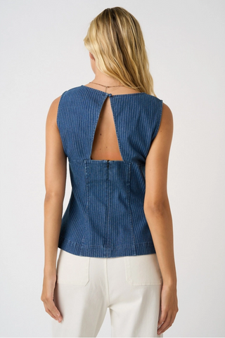 A woman viewed from the back wearing a sleeveless blue denim-style top with a cutout back and white pants, against a plain light gray studio background.